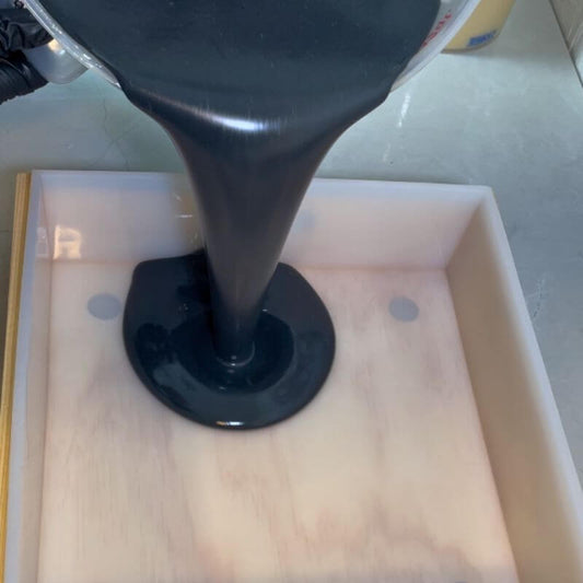 Handmade soap making at Sudsverse: pouring tea tree and charcoal soap mixture into a mold with natural ingredients like olive oil and peppermint leaves
