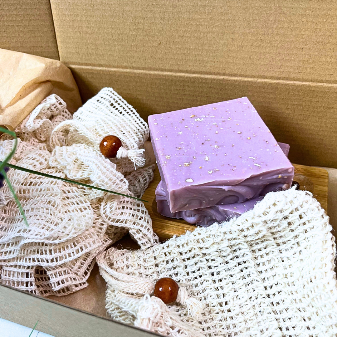 A self care starter set from Sudsverse showing 2 bars of Lavender Oatmeal Natural Handmade Soaps on a bamboo soap dish, a soapsaver bag and a ramie mesh pouf in an eco friendly gift box.