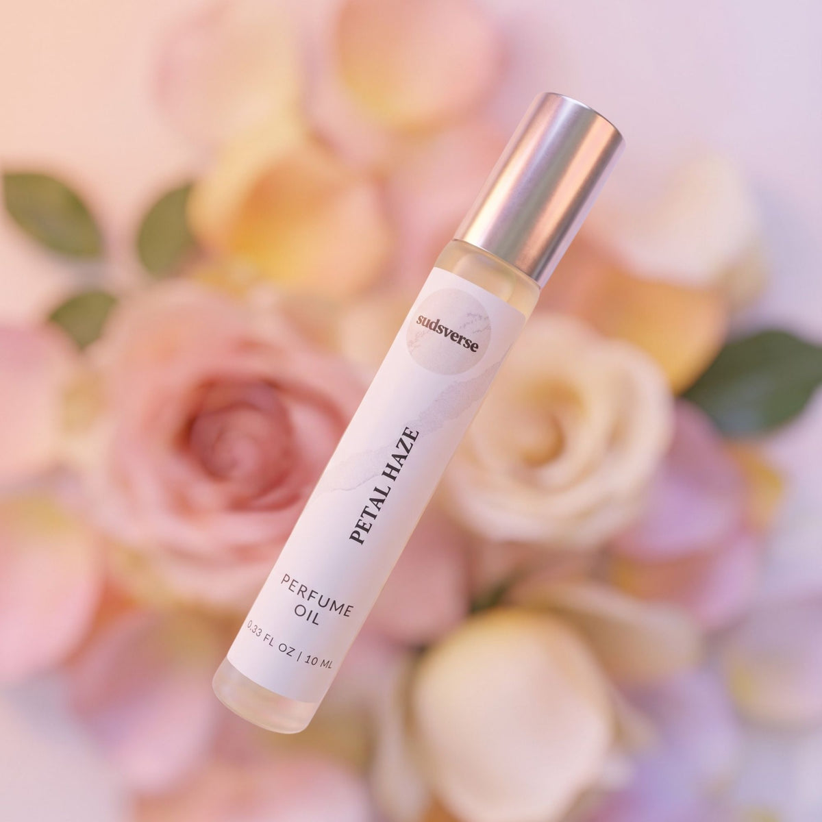 A 10ml bottle of 'Petal Haze' perfume oil by the brand 'sudsverse' is shown diagonally against a soft-focus background of pink and yellow roses. The bottle has a white label with black text and a metallic silver cap.