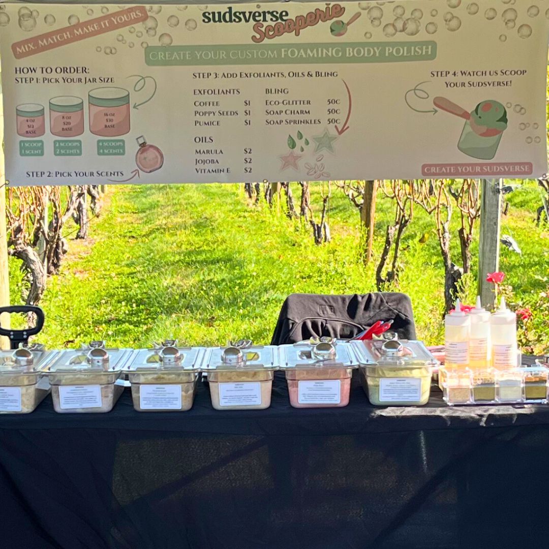 Sudsverse Scooperie showing a table with containers and a sign for creating custom foaming body polish outdoors.